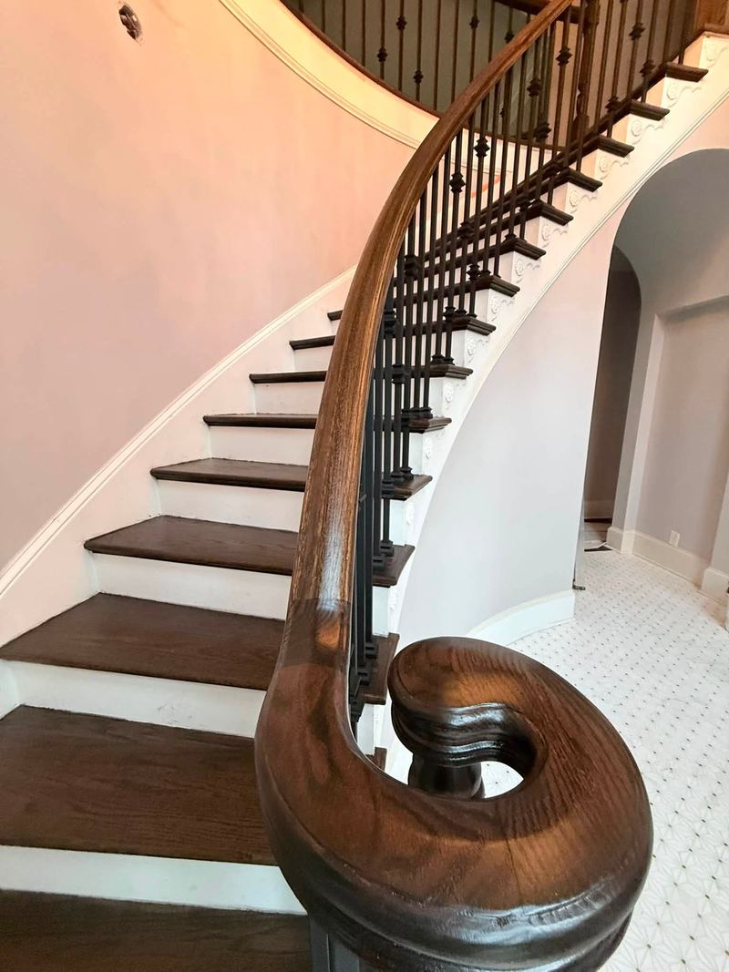 Curved staircase with dark stained treads, white risers, iron balusters and a carved handrail volute